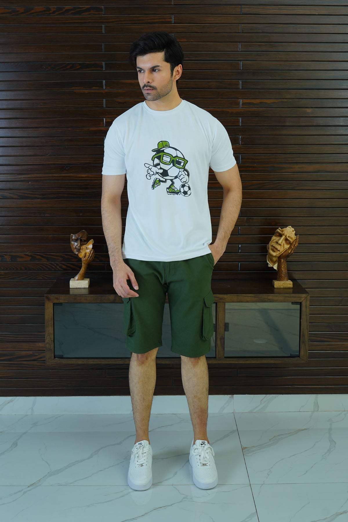 Man wearing a white t-shirt with a graphic design and green shorts standing against a wooden paneled wall.