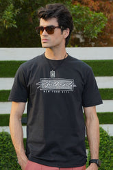 A male model wearing sunglasses and a black t-shirt with a bold graphic print on the front, standing in a garden setting.