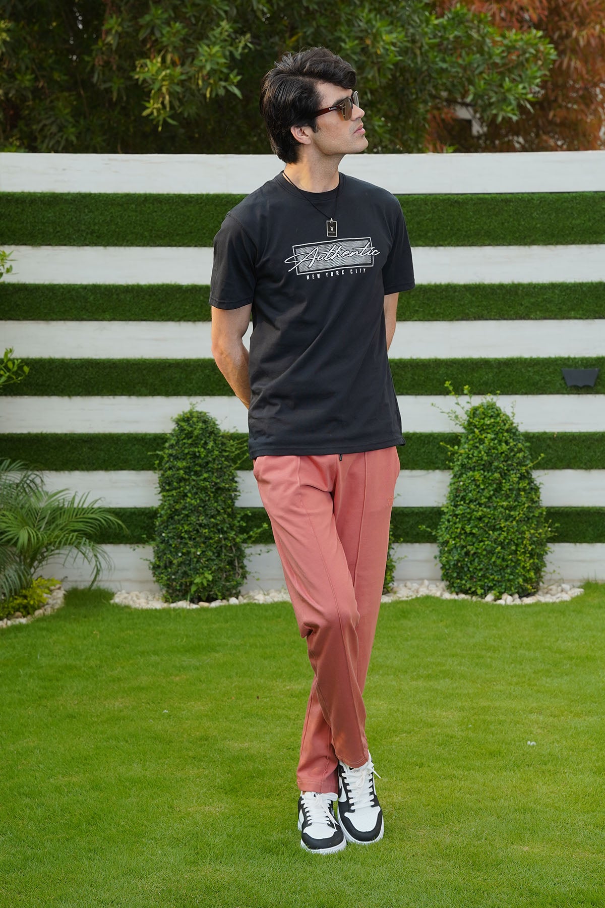 Man wearing a black t-shirt and pink pants standing in a grassy area with a white fence and greenery in the background.