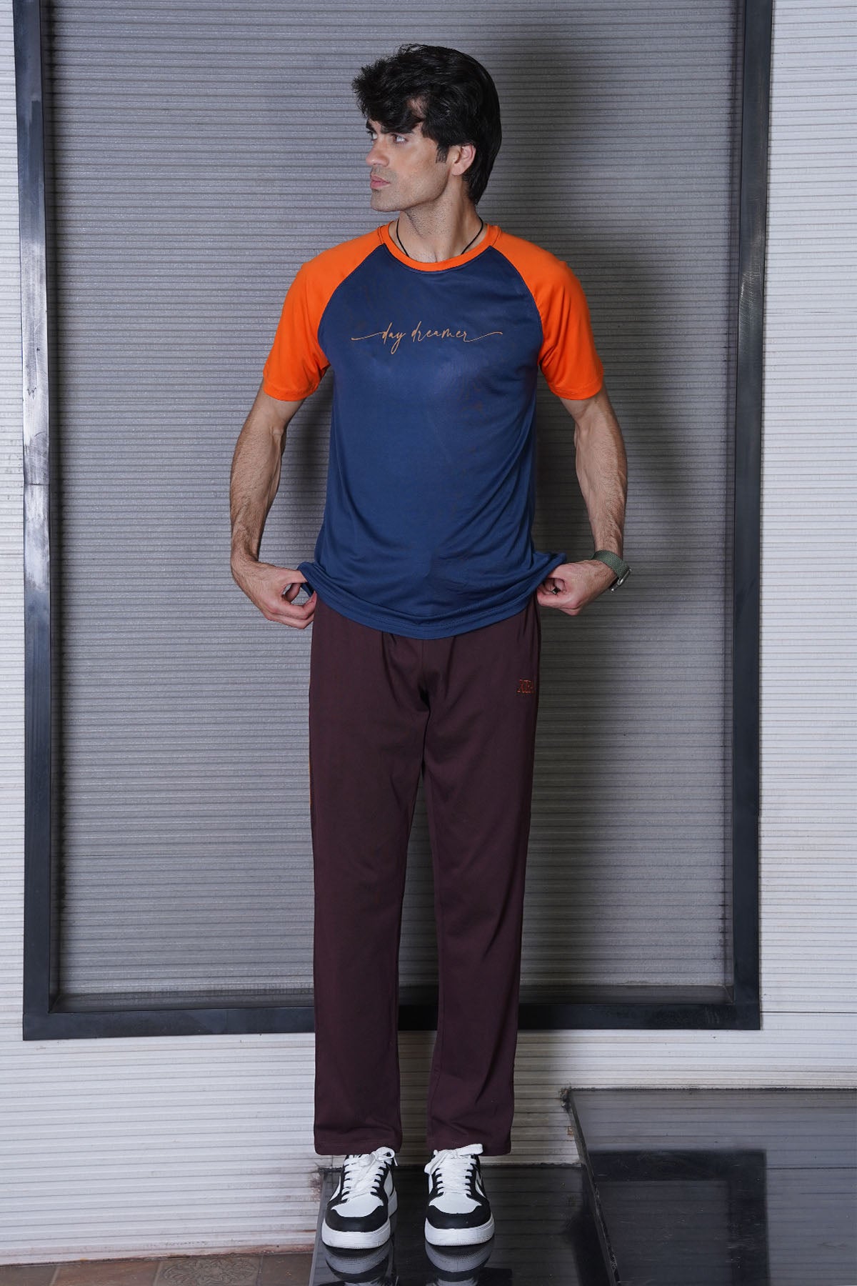 Man wearing a blue and orange shirt with maroon pants standing against a white wall.