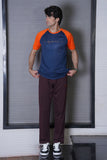 Man wearing a blue and orange shirt with maroon pants standing against a white wall.