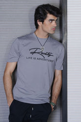 Man wearing a gray t-shirt with text, standing against a metallic wall.