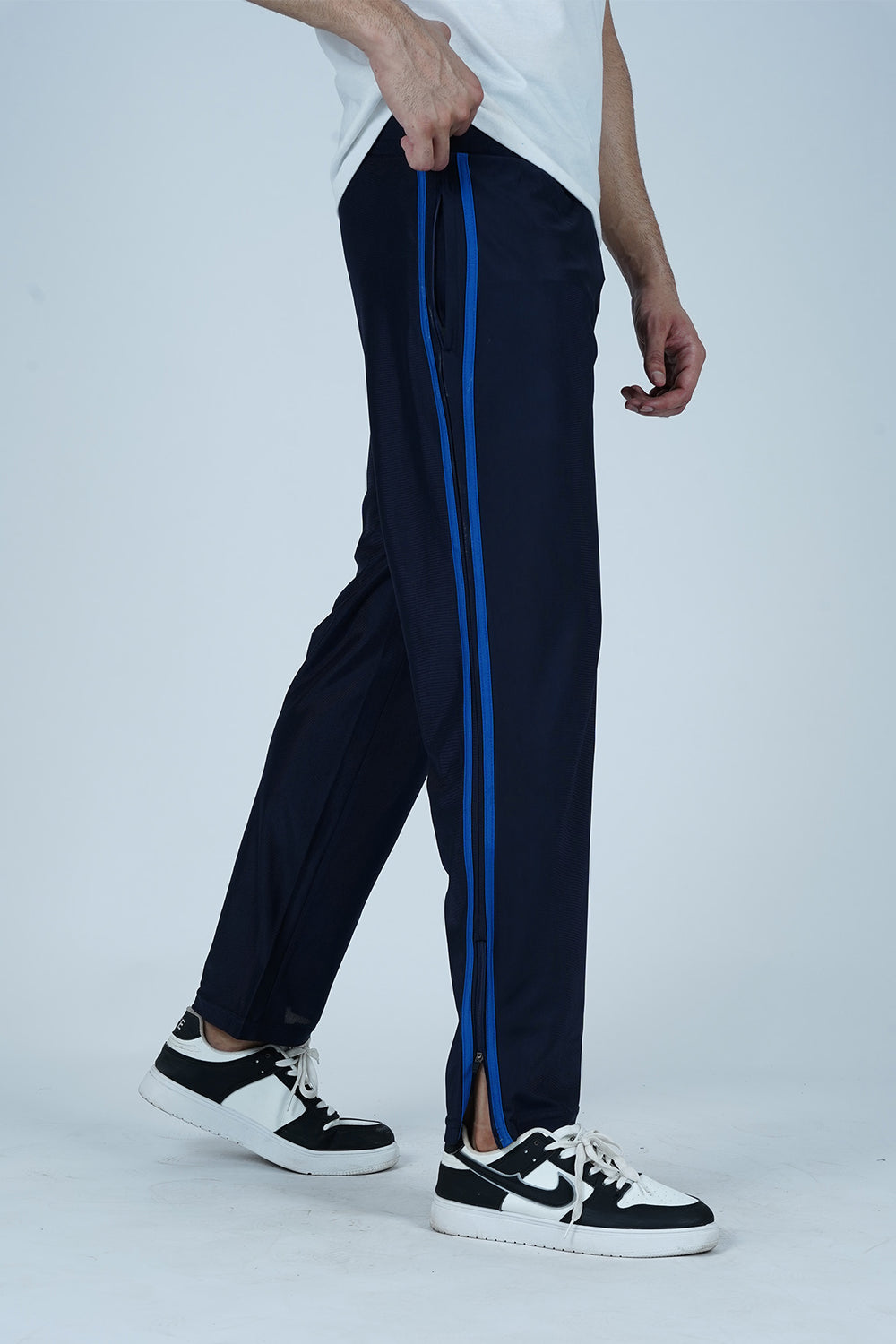 A person standing side-on, wearing navy track pants with blue contrast side stripes, a white t-shirt, and white sneakers.
