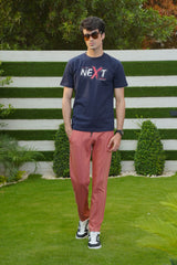 A person wearing a navy blue graphic t-shirt with the word 'NEXT' printed on the front, paired with red pants and standing in front of a white fence.