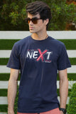 A person wearing a navy blue graphic t-shirt with the word 'NEXT' printed on the front, paired with red pants and standing in front of a white fence.