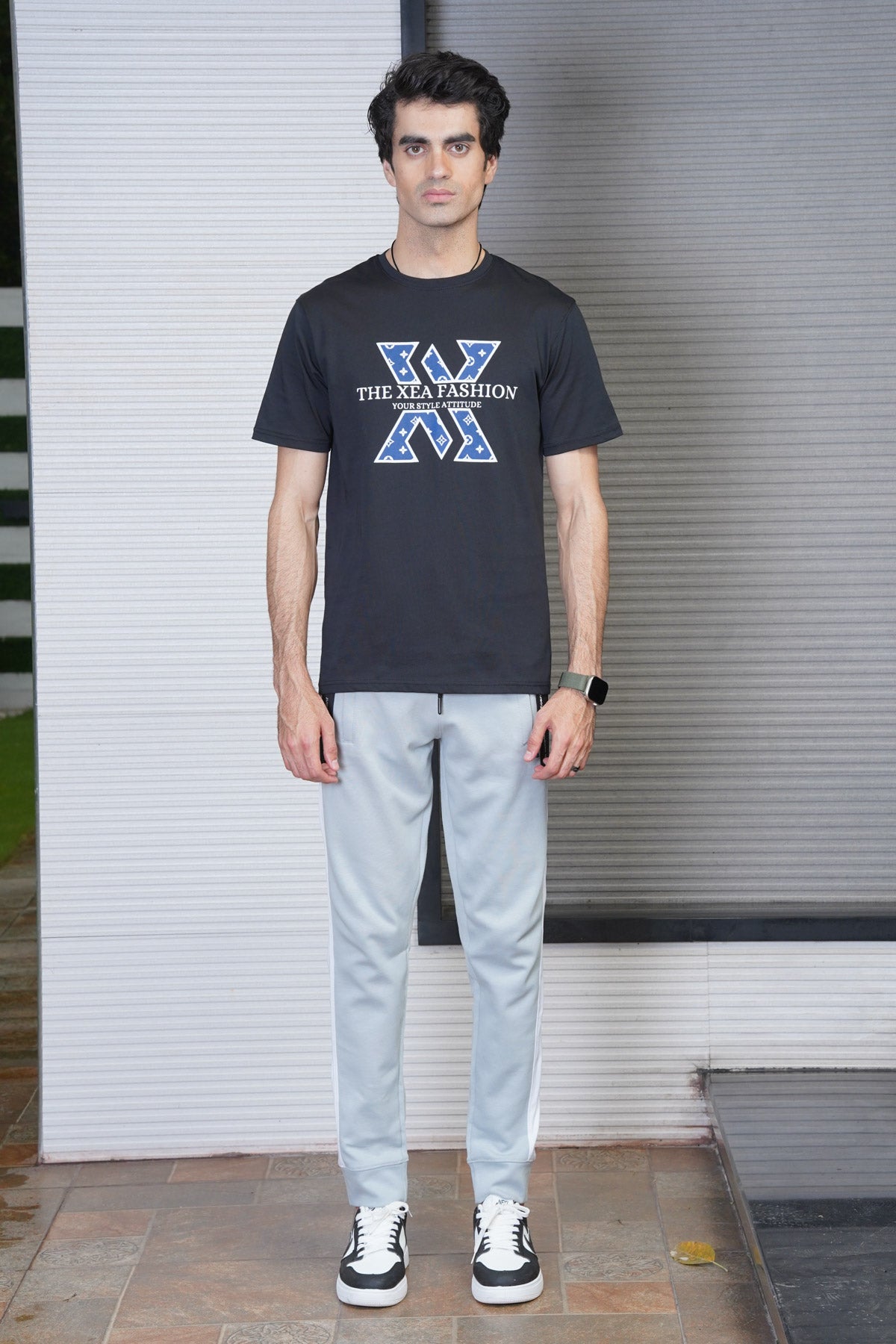 A man standing in front of a white wall wearing a black crew neck T-shirt with a graphic text design, light gray trousers, and black and white sneakers.