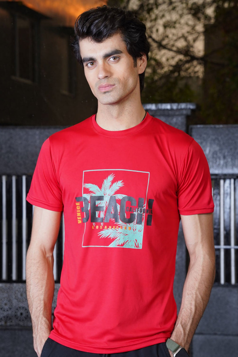 A male model wearing a red Venice Beach graphic T-shirt with a palm tree and text design on the front.