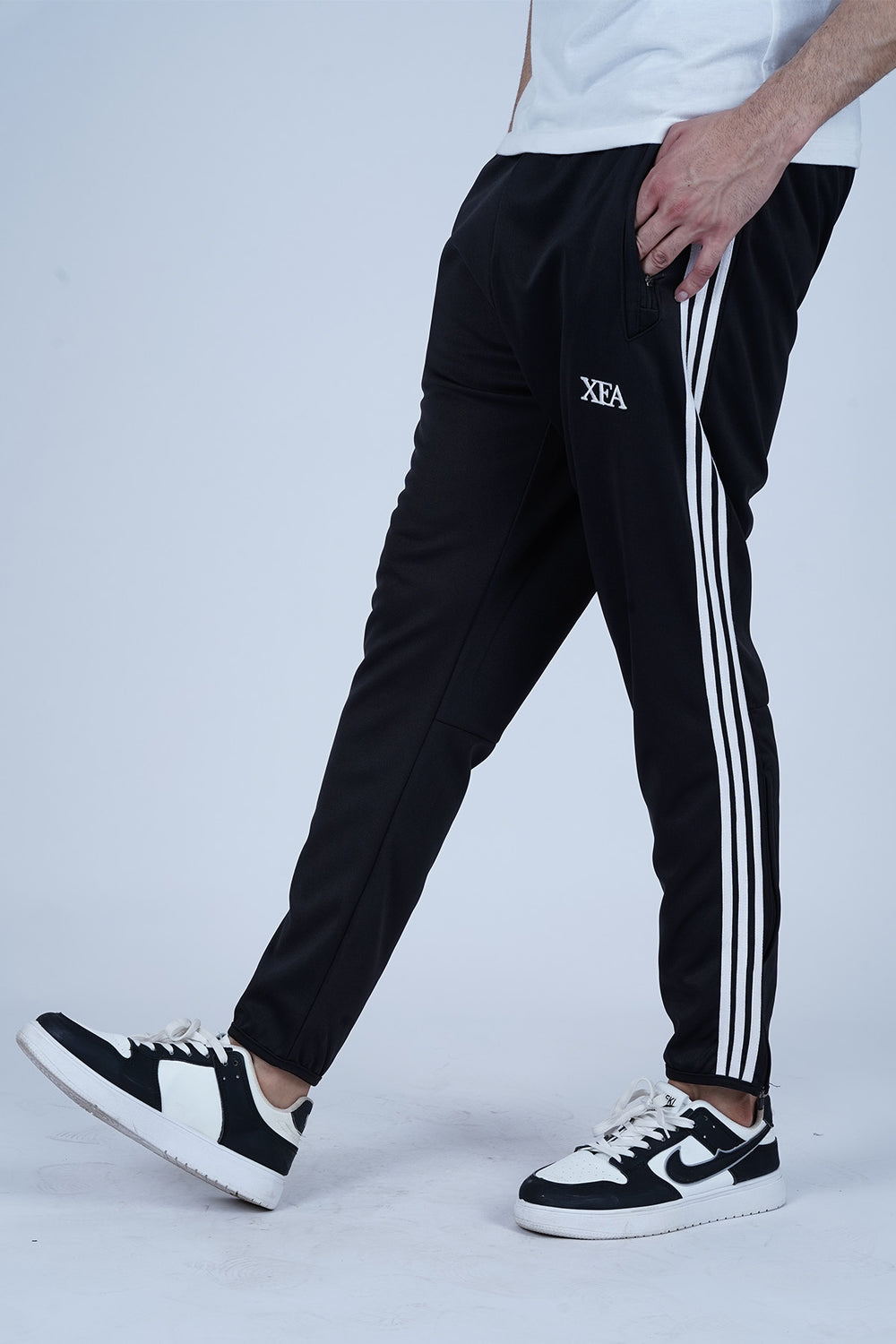 A man wearing black trouser/pants with white stripes on the side, a white t-shirt, and white sneakers. The image shows the pants in a vertical orientation.