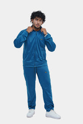 A man wearing a blue tracksuit, consisting of a hooded jacket and pants, standing against a white background.