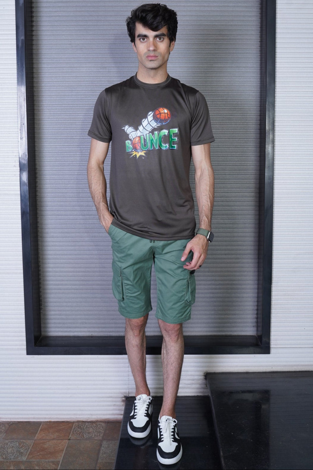 Man wearing a gray t-shirt with colorful graphics and green shorts standing in front of a window.
