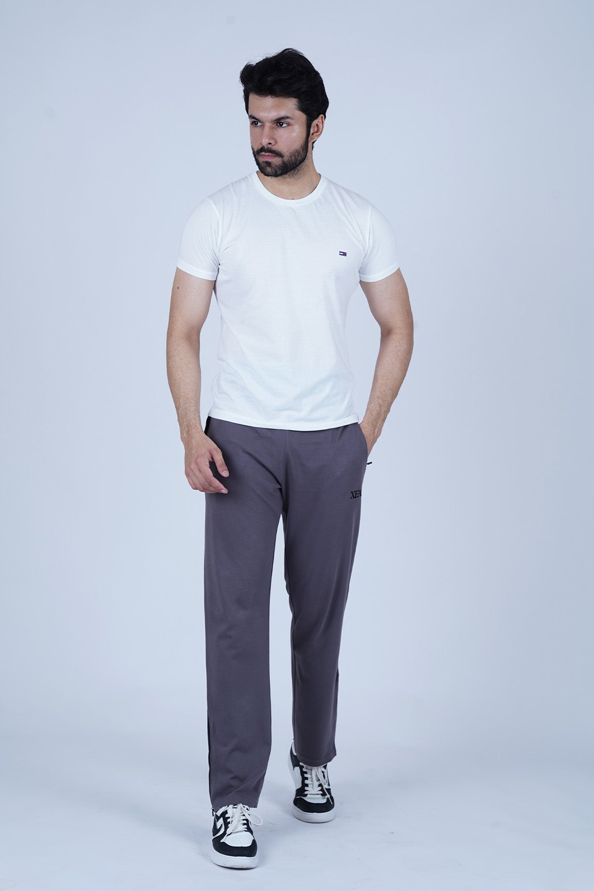 A man wearing a white crew neck t-shirt and gray track pants with a zipper and pockets, standing against a plain background.