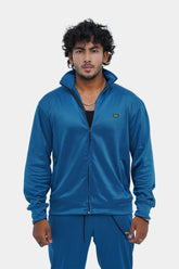 A man wearing a blue tracksuit, consisting of a hooded jacket and pants, standing against a white background.