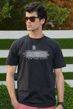 A male model wearing sunglasses and a black t-shirt with a bold graphic print on the front, standing in a garden setting.