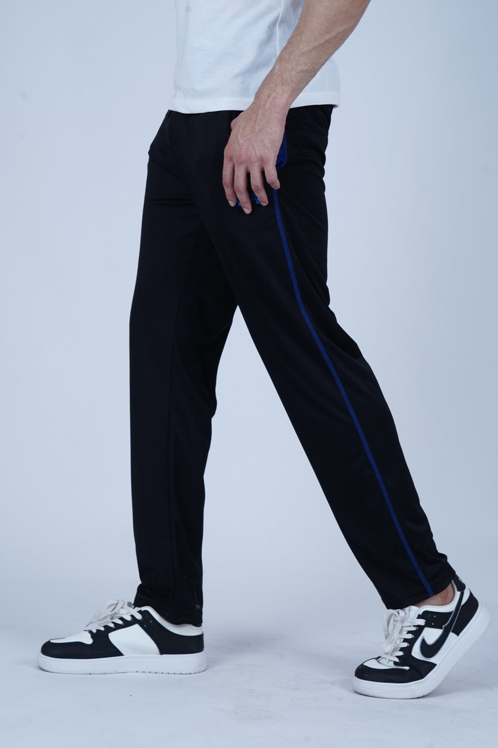 A man wearing black track pants with blue stitching and a white t-shirt, standing against a blue background.