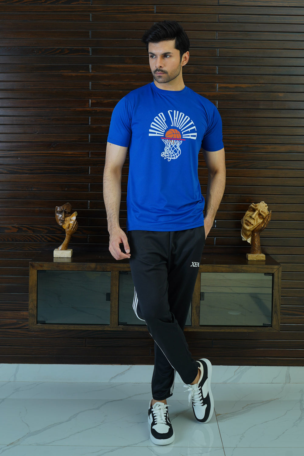 A man wearing a royal blue graphic t-shirt with a basketball design, paired with black pants and white sneakers, standing in front of a wooden backdrop with two golden trophies on display.