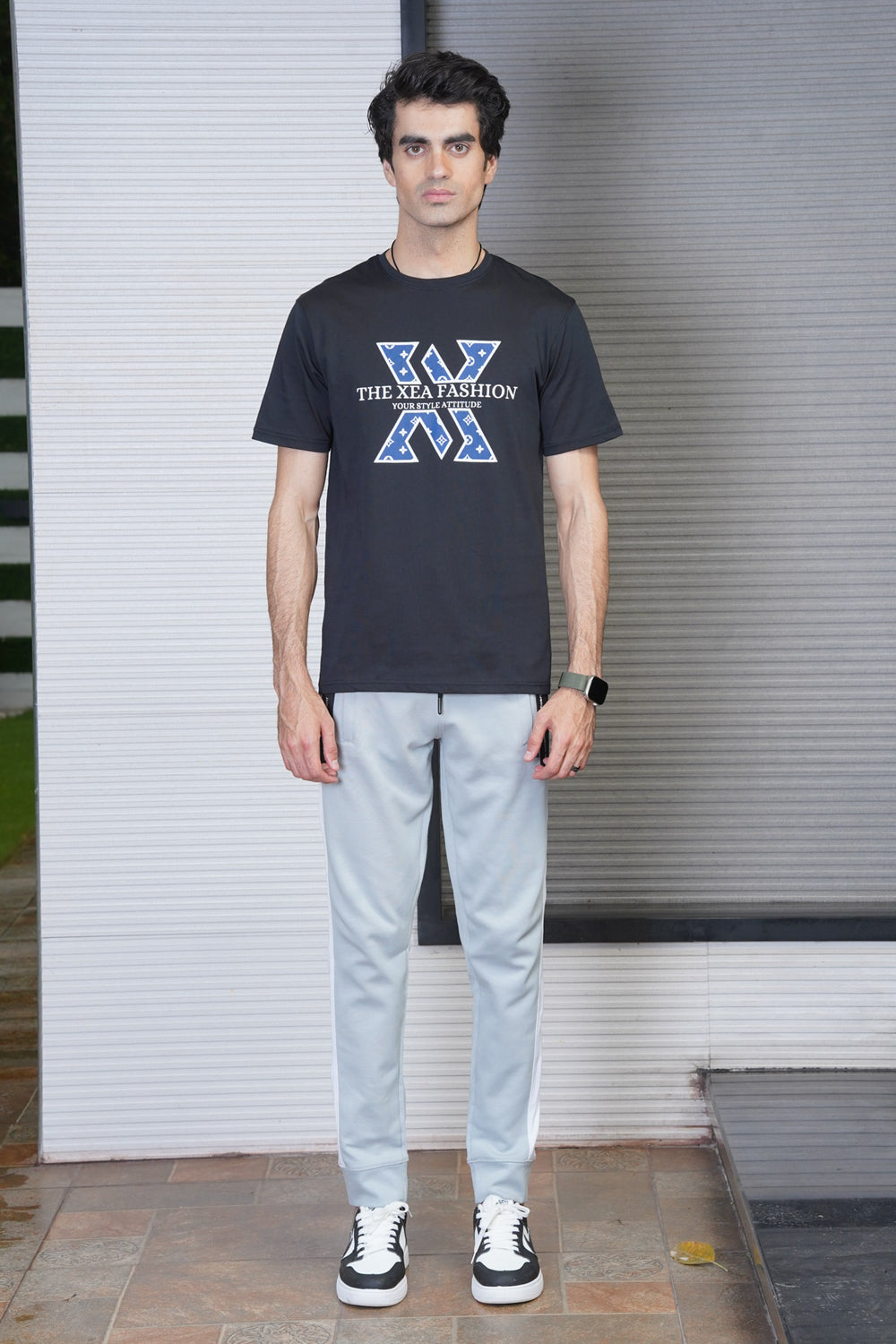 A man standing in front of a white wall wearing a black crew neck T-shirt with a graphic text design, light gray trousers, and black and white sneakers.