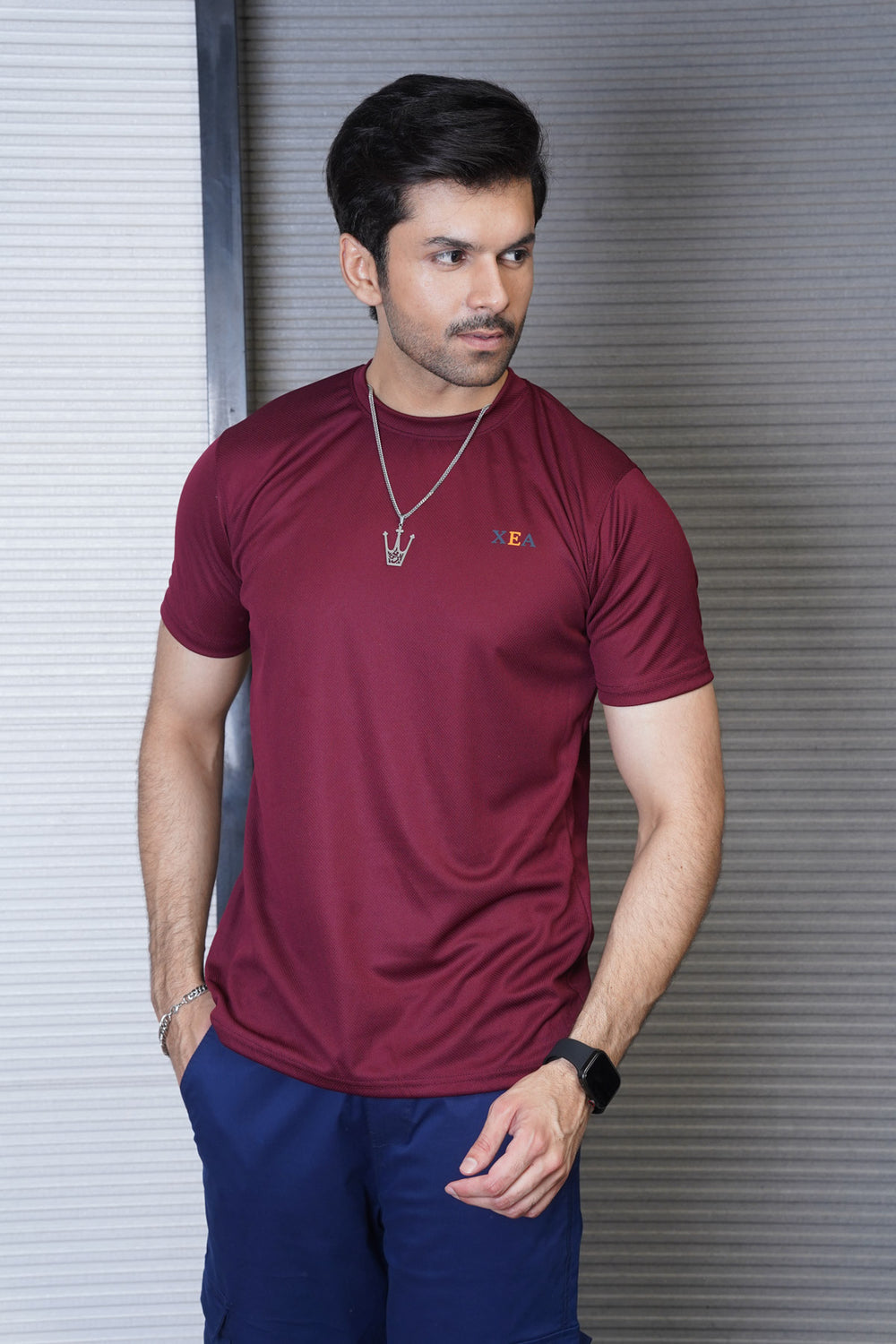 A man wearing a maroon crew neck t-shirt and blue trousers. The t-shirt has a small graphic on the left chest area.