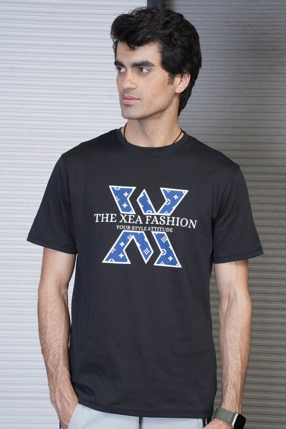 A male model wearing a black crew neck t-shirt with a graphic design in the center, standing in front of a metallic background.