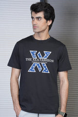 A male model wearing a black crew neck t-shirt with a graphic design in the center, standing in front of a metallic background.