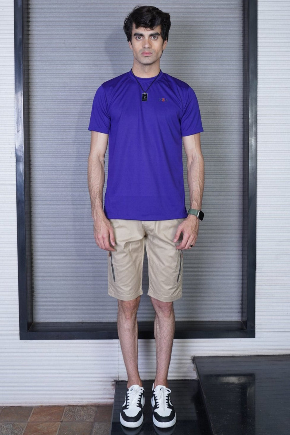 A man standing, wearing a purple crew neck t-shirt and beige shorts with white sneakers.